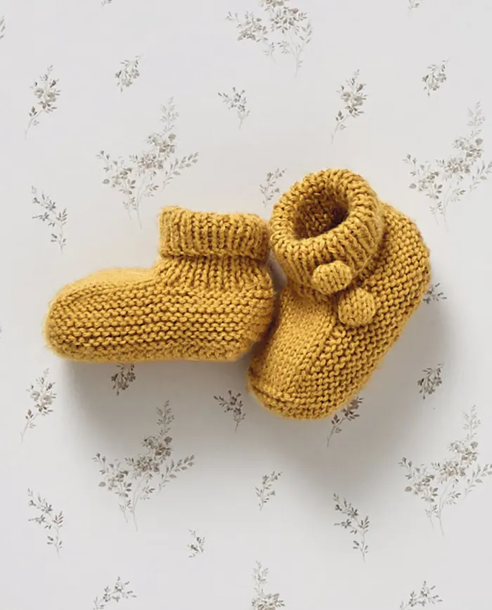 Botins de bebé em malha mostarda com pompons sobre fundo branco com padrão de ramos.