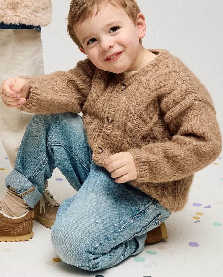 Criança com camisola castanha de malha e jeans sentado no chão com confetes coloridos