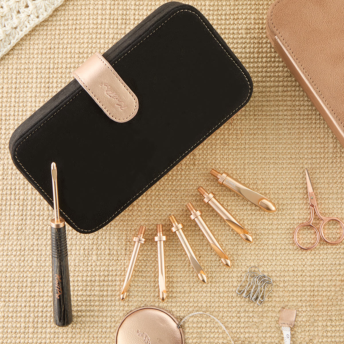 Estojo preto e acessórios de costura dourados e rosa sobre tecido bege