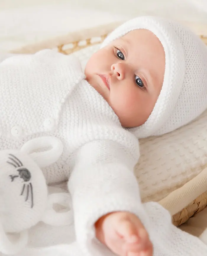 Bebé com roupa de malha branca e gorro, deitado numa almofada creme.