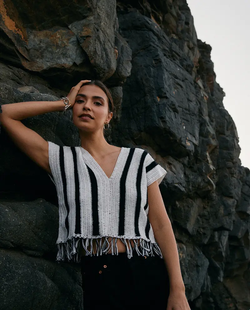 Mulher vestindo top de malha branco com listra preta, decote V e franjas, em cenário rochoso ao ar livre