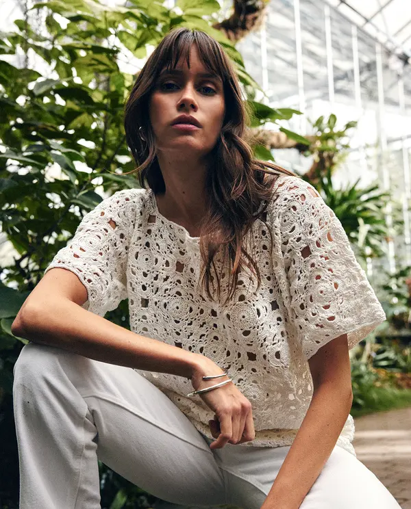 Mulher com blusa branca de malha rendada quadrada e calças claras sentada em ambiente com plantas