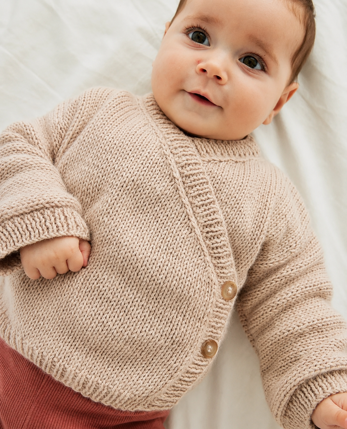 Bebé com camisola bege de malha e calças rosa-encarnado deitado sobre tecido branco