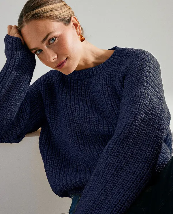 Mulher com camisola azul escura de malha e brincos dourados, fundo cinza claro