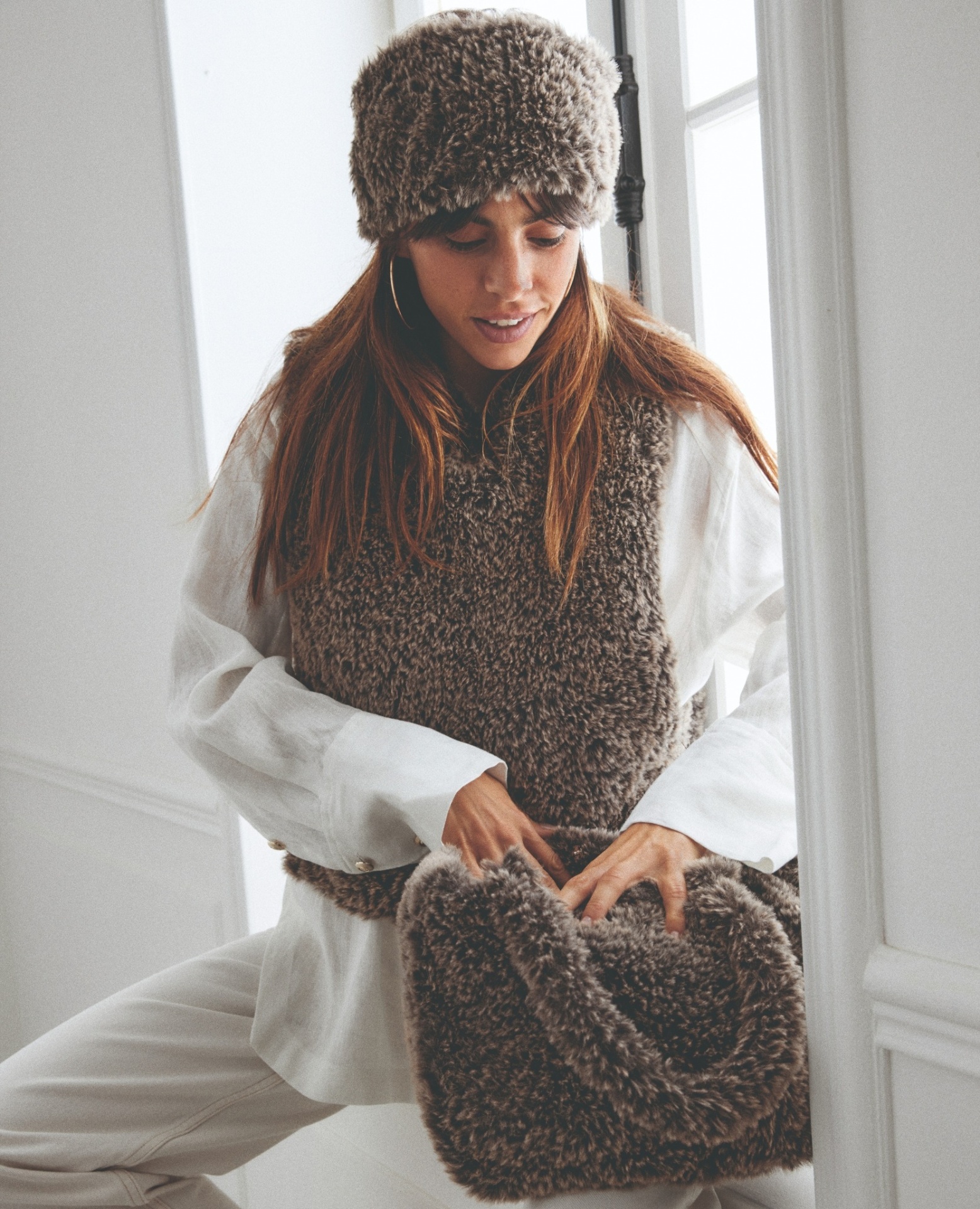 Mulher com colete e gorro felpudo castanhos e camisa branca, segurando bolsa castanha felpuda