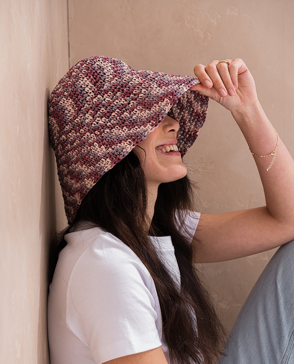 Mulher a usar chapéu tricotado colorido e roupa branca, encostada a parede bege.