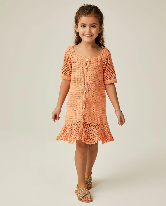 Menina com vestido laranja de crochet e sandálias coloridas em fundo neutro
