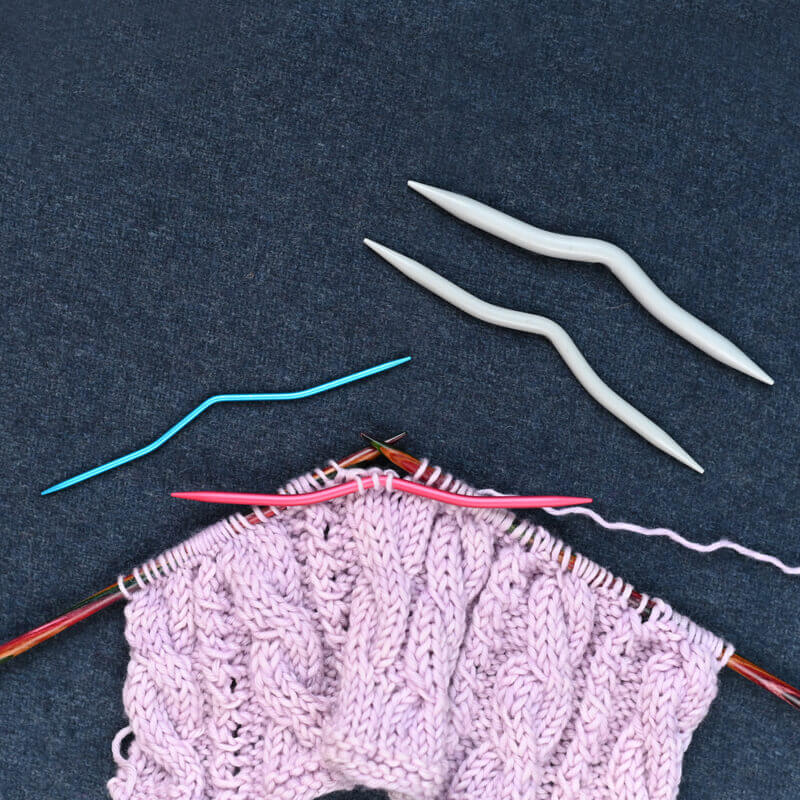 Trabalho de tricot rosa com agulhas de madeira, plásticas cinzentas e uma agulha azul sobre tecido azul