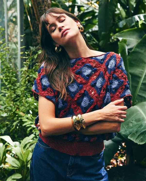 Mulher com camisola crochet azul e vermelha, calças azuis e acessórios dourados num ambiente verde