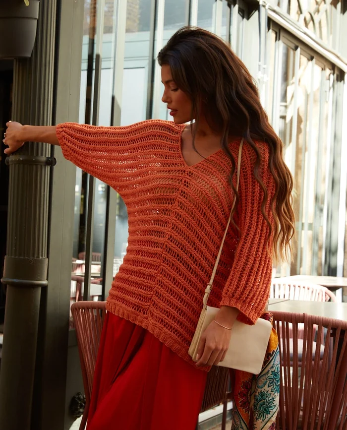 Mulher com camisola de malha laranja e saia vermelha, segurando carteira bege junto a um poste verde