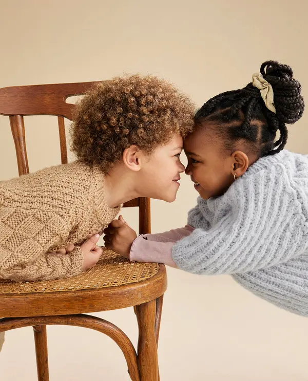 Crianças em camisolas de malha tricotada encostadas de testa, uma sentada numa cadeira de madeira castanha.