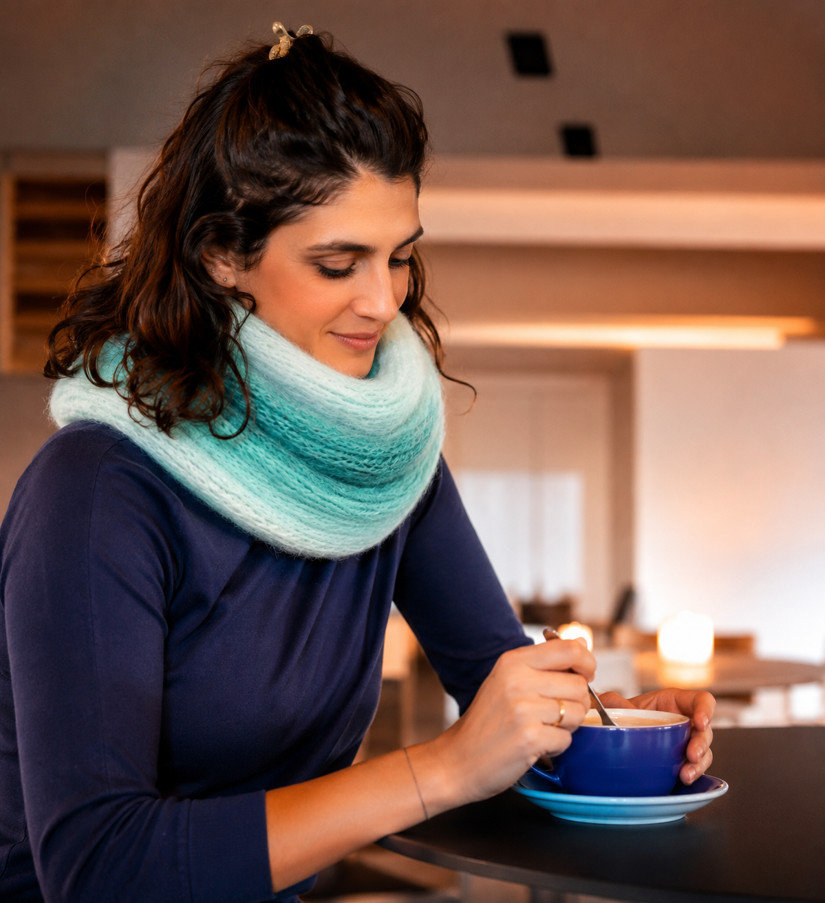 Cachecol azul claro e turquesa em malha grossa numa mulher com blusa azul escura mexendo bebida quente numa chávena azul.
