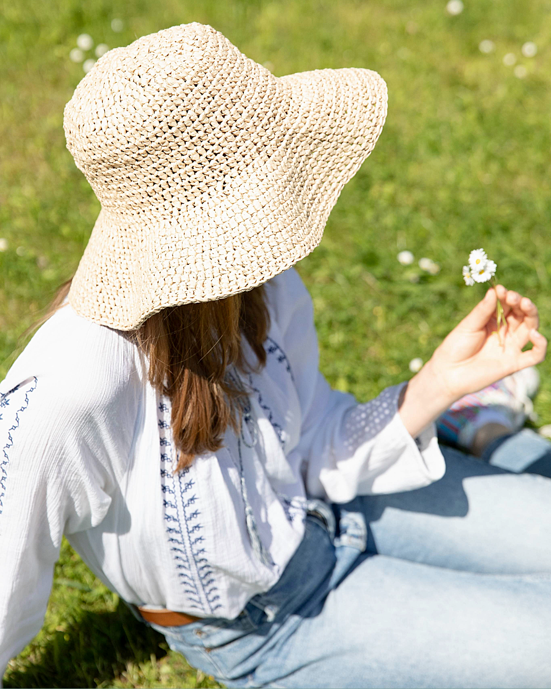 Pessoa com chapéu de palha e roupa casual sentada na relva segurando flores.