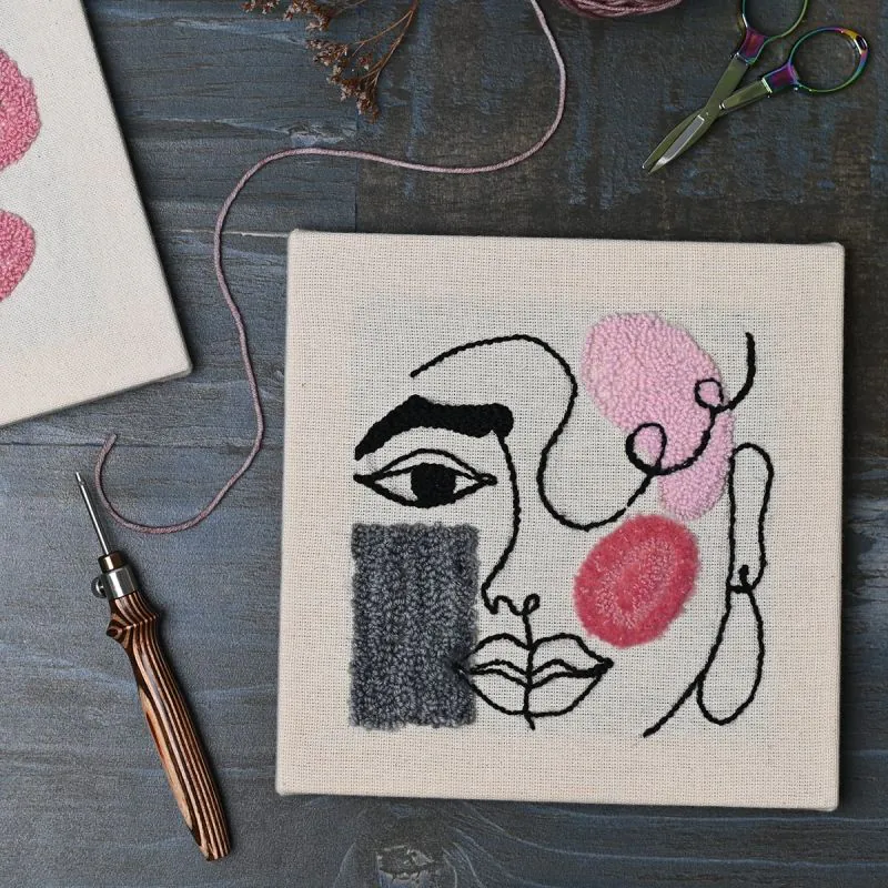 Tela branca bordada com rosto estilizado em linhas pretas e manchas rosa e cinza