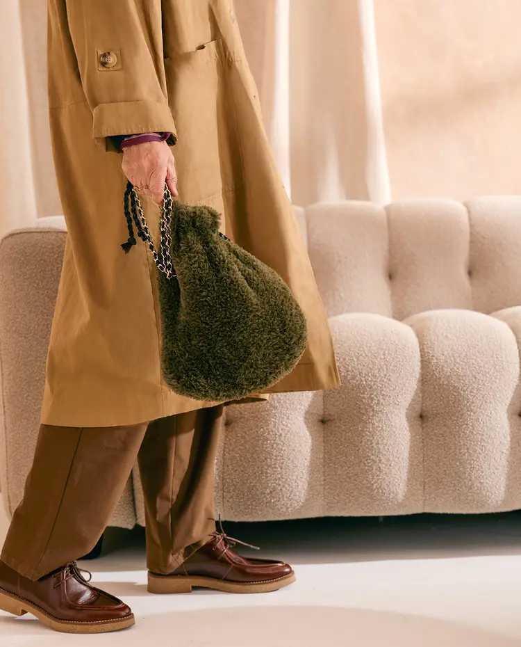Pessoa com mala verde felpuda e roupa castanha junto a sofá bege