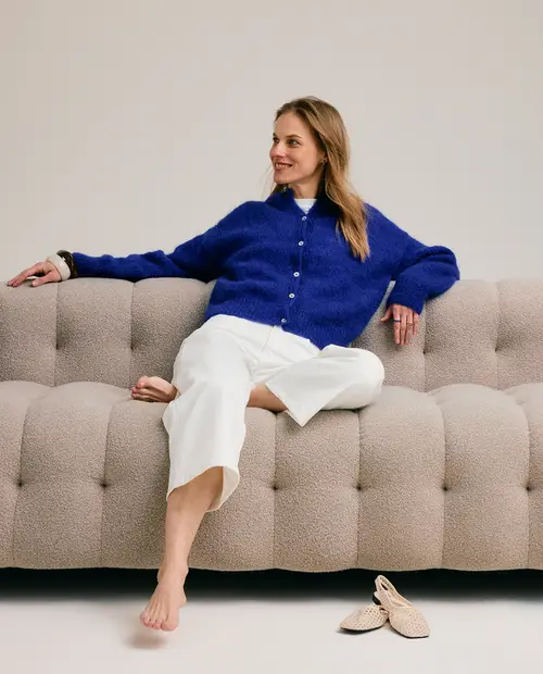 Mulher sentada num sofá cinza claro usando camisola azul e calças brancas com sapatos bege ao lado