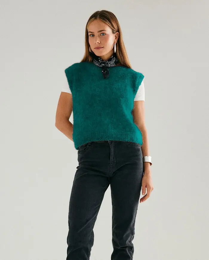 Mulher com camisola de malha verde turquesa sem mangas, lenço preto ao pescoço e calças de ganga pretas num fundo branco
