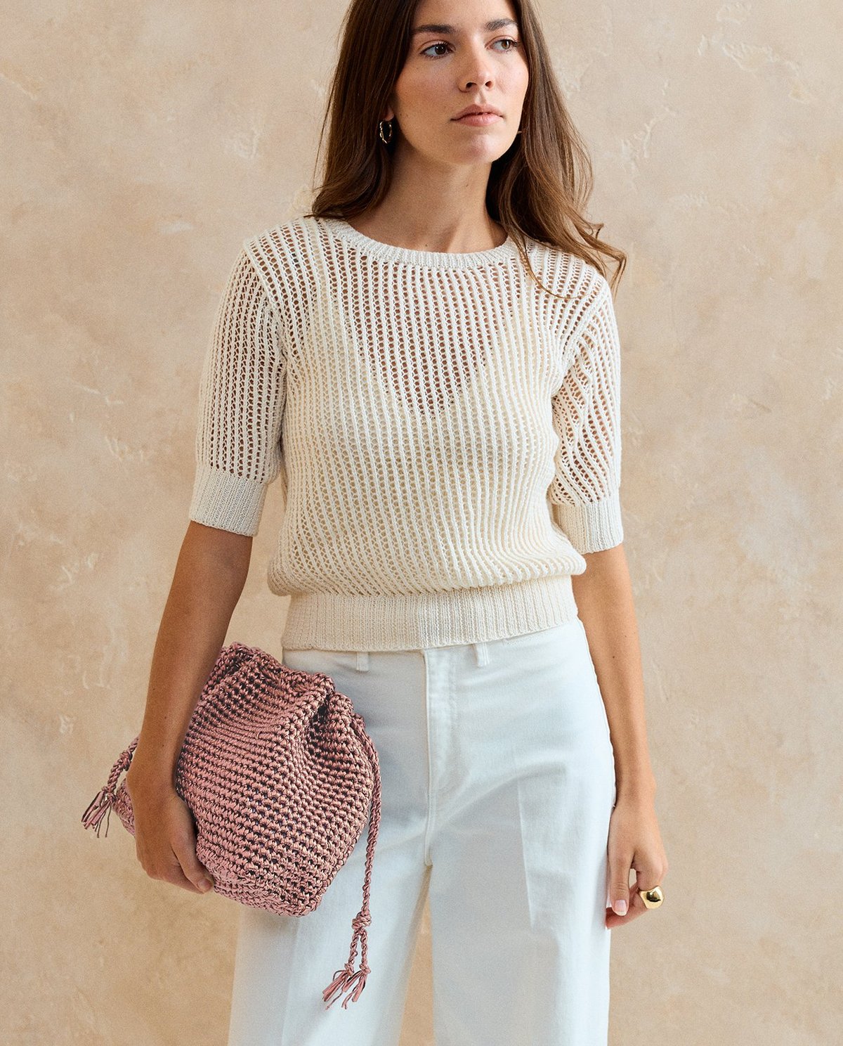 Mulher com camisola branca de malha e calças brancas segurando mala rosa trançada