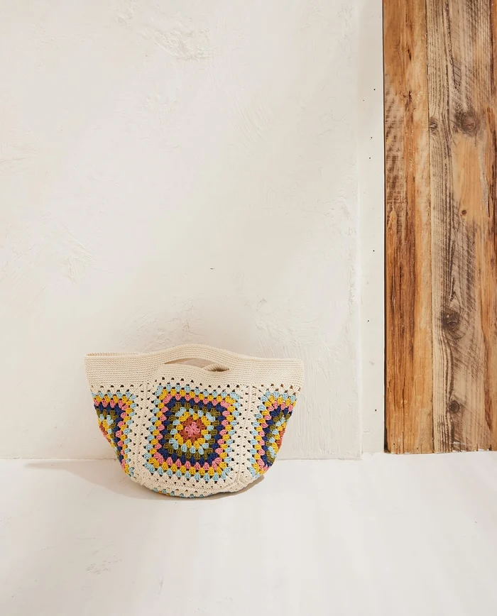 Bolsa de croché creme com padrões coloridos encostada numa parede branca e madeira