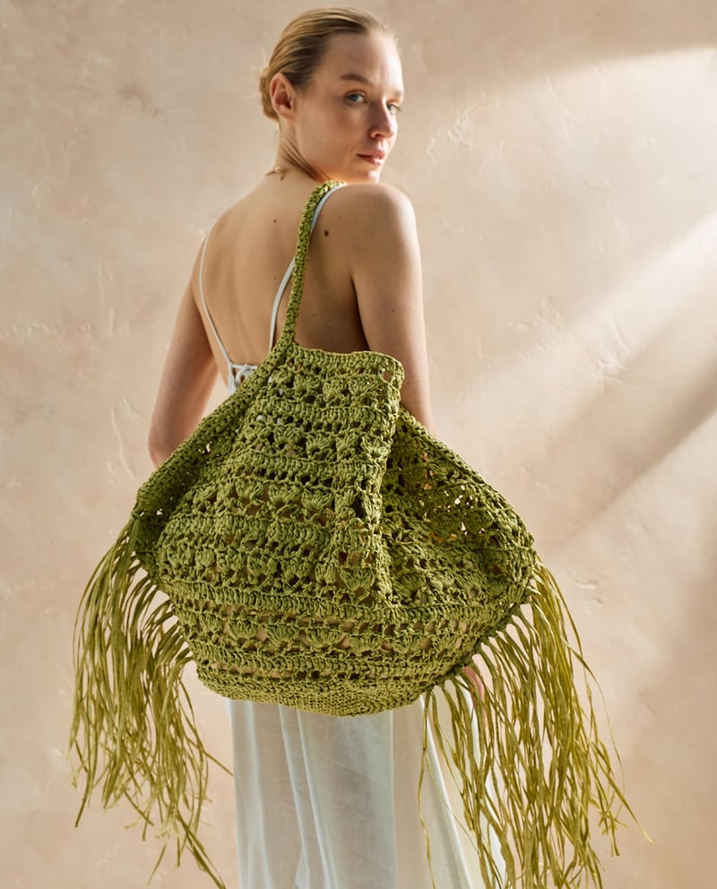 Mala feminina verde em crochet com franjas usada por mulher com vestido branco.