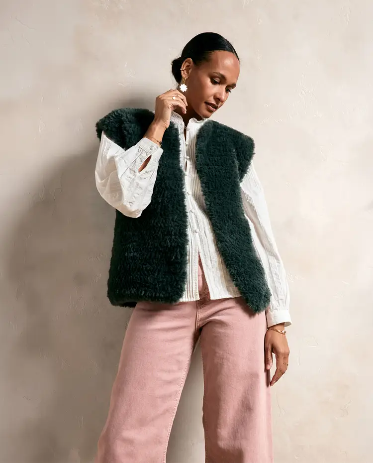 Mulher vestindo colete felpudo verde-escuro, camisa branca e calças rosa claro contra parede bege.
