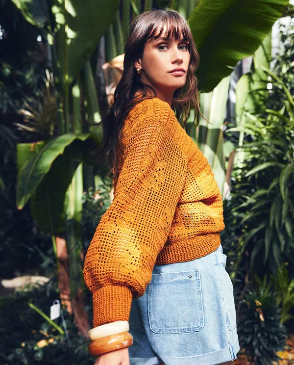 Mulher com camisola de malha laranja e calções de ganga em ambiente com plantas tropicais