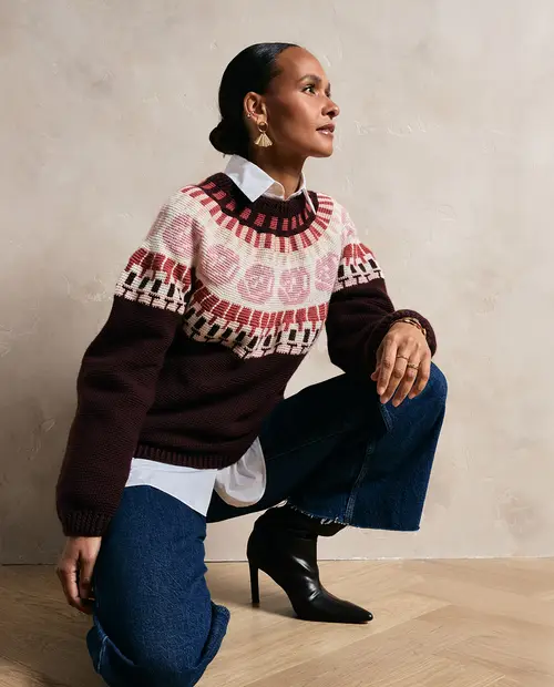 Mulher com sweater de malha castanho com padrão rosa e branco, camisa branca, jeans azuis e botins pretos de salto