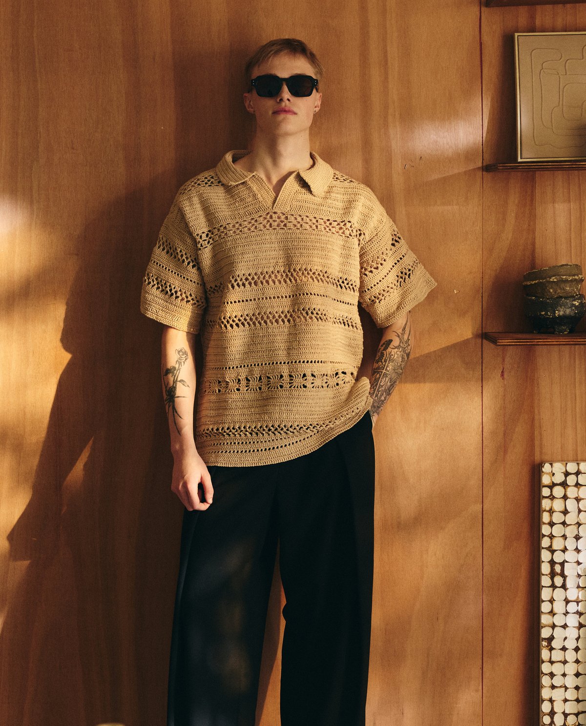 Pessoa com tshirt de malha bege, calças pretas e óculos de sol contra parede de madeira em interior