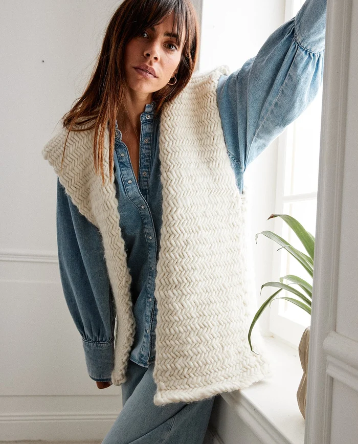 Mulher com colete creme de malha grossa e camisa jeans azul clara junto a janela