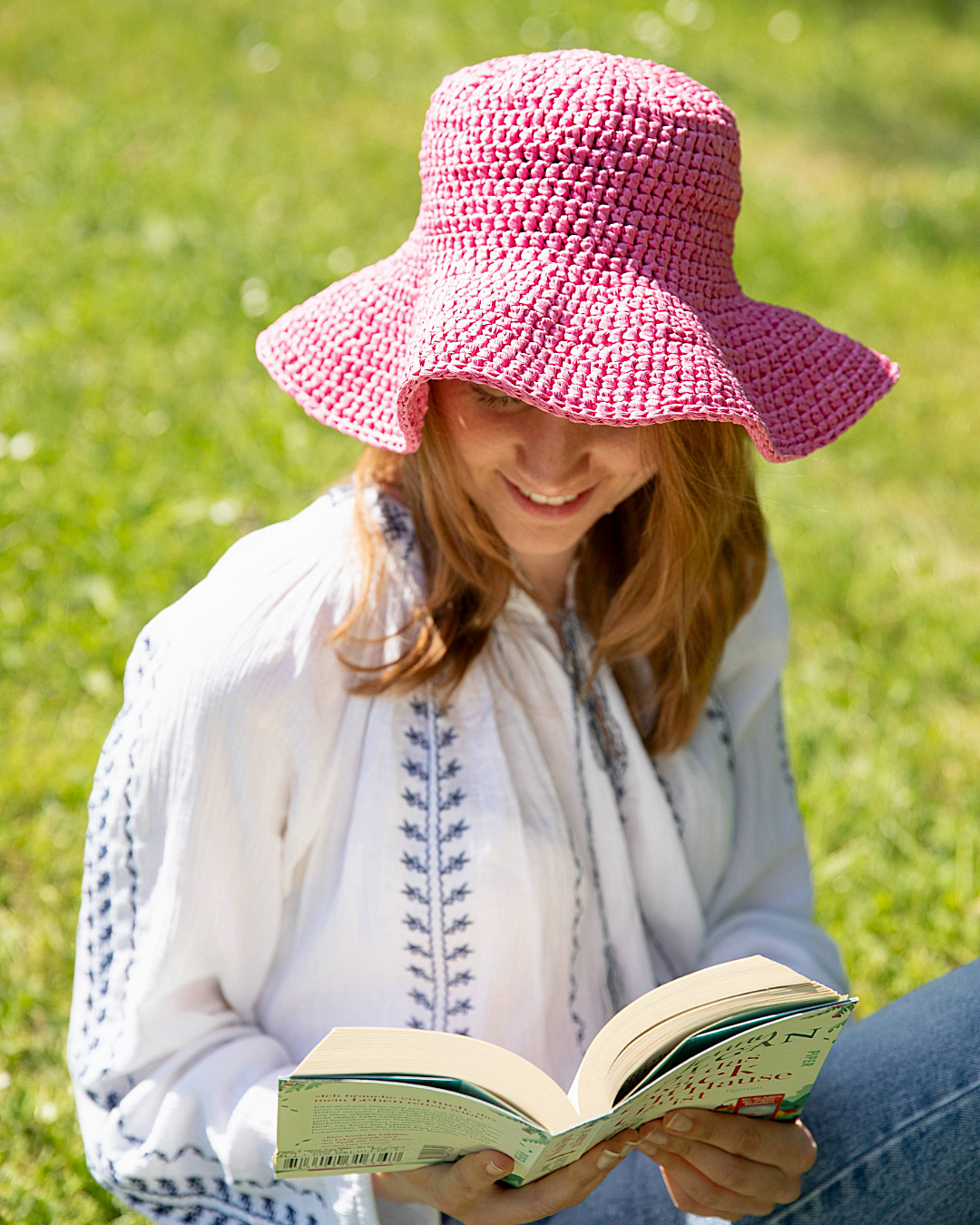 Mulher com chapéu rosa e blusa branca a ler livro na relva