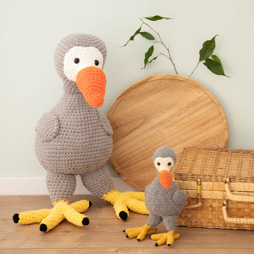 Duas peluches de crochet aves cinzentas bicos laranja e pés amarelos junto a mala de vime e prato de madeira