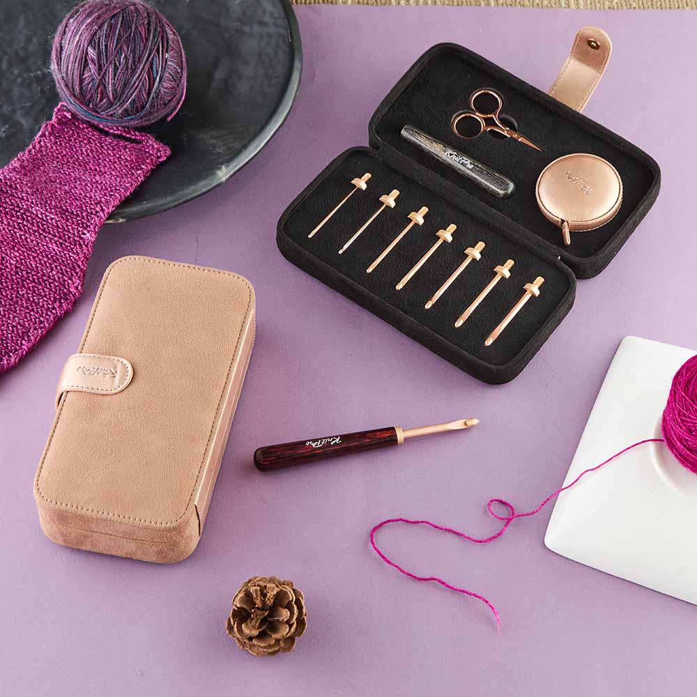 Conjunto de agulhas de tricô com estojo bege aberto e fios de lã roxos sobre mesa lilás