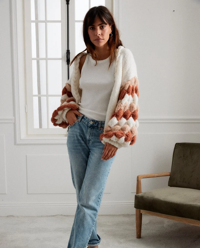 Mulher com cardigan de malha felpuda branca, bege e castanha, camisola branca e jeans azuis, em sala interior