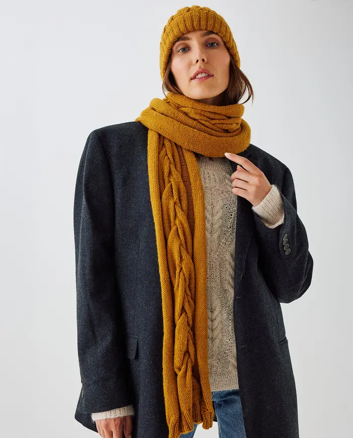 Mulher com camisola bege, casaco cinza, cachecol e gorro amarelo mostarda no fundo branco