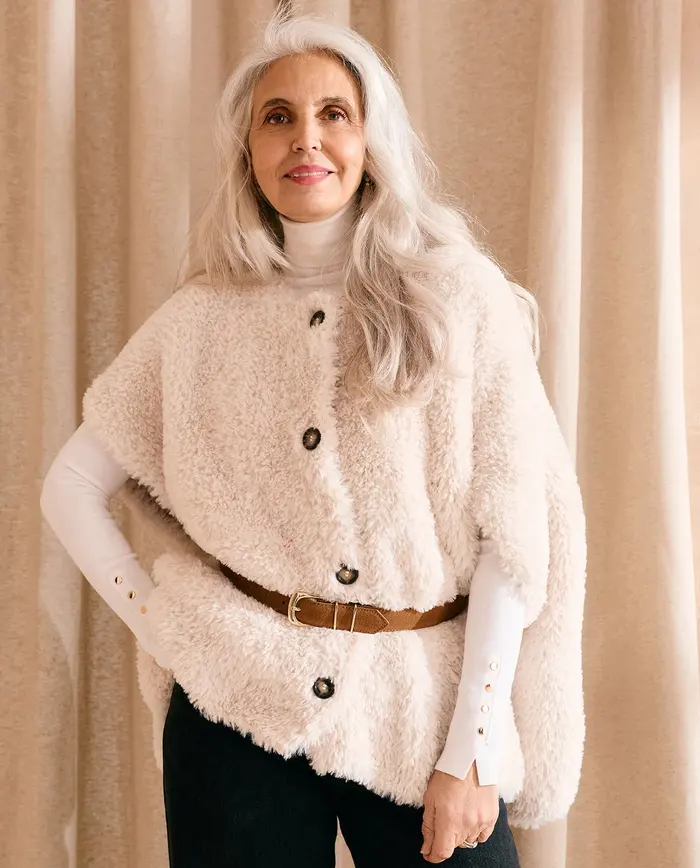 Mulher com capa felpuda creme, botões e cinto castanho em fundo com cortinados creme