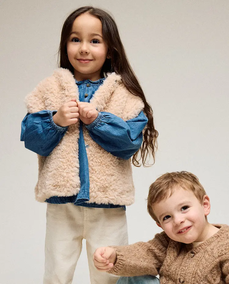 Duas crianças vestidas com roupa de inverno, uma com colete felpudo e outra com camisola de malha.