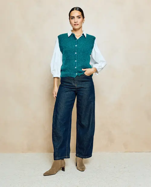 Mulher com camisola verde, camisa branca, calças de ganga e botas castanhas contra parede bege