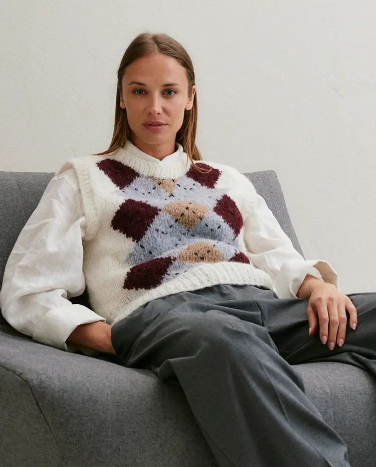 Mulher sentada num sofá cinzento a usar colete de malha branco com padrão colorido e camisola branca