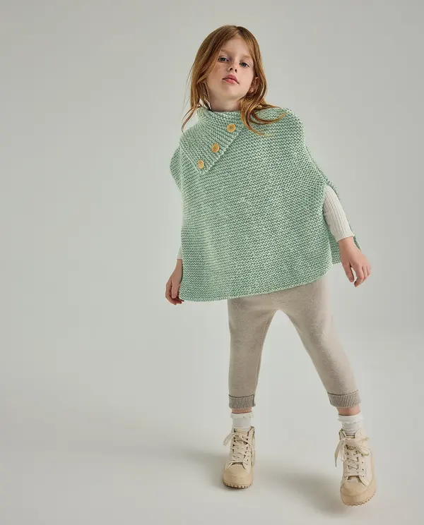 Criança com capa de malha verde água, calças bege e sapatilhas bege em fundo cinza