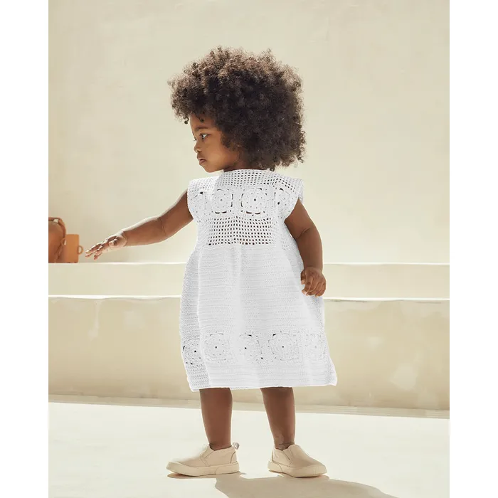 Criança vestindo vestido branco de croché com padrão floral e calçado bege em fundo neutro claro