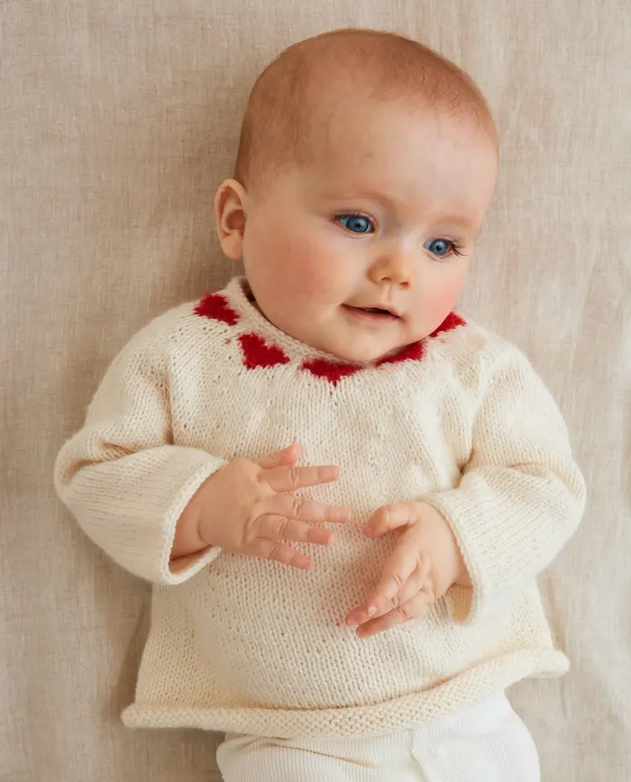 Bebé deitado com camisola de malha branca com corações vermelhos no pescoço.