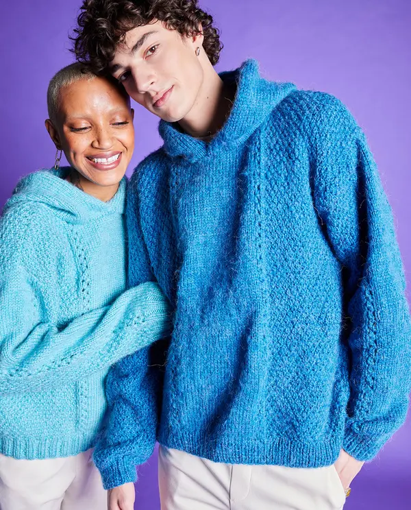 Dois modelos com camisolas de malha azul com capuz e textura trançada frente a fundo púrpura