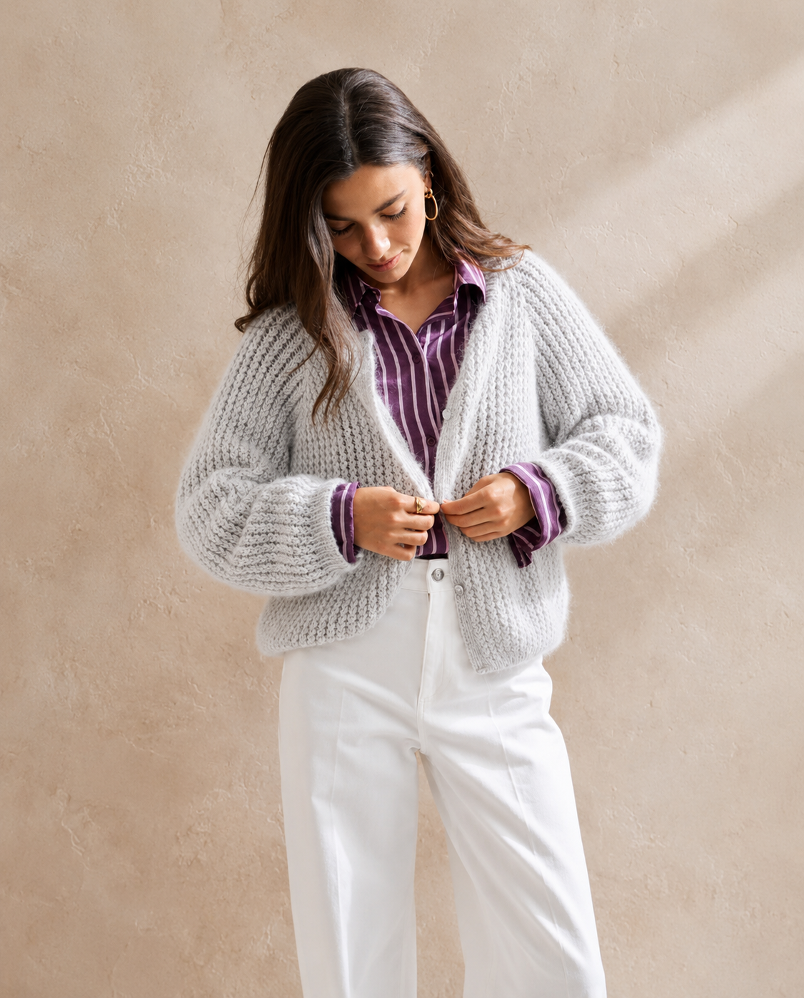 Mulher a vestir cardigan cinzento claro de malha grossa, camisa aos riscos roxa e calças brancas.