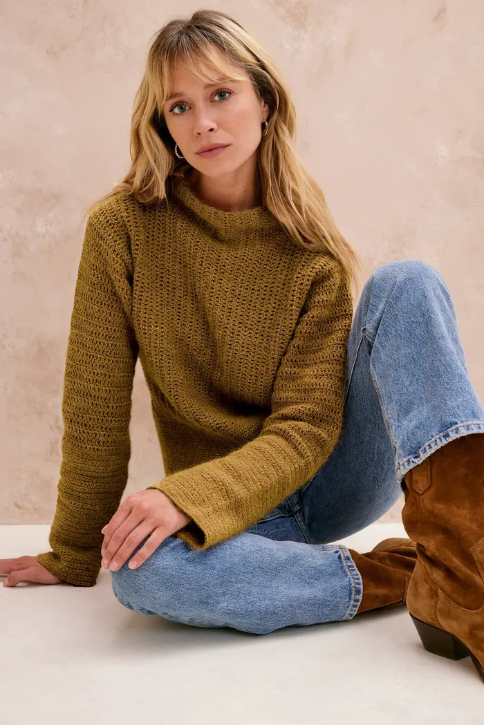 Mulher com camisola castanha, jeans azuis e botas castanhas sentada em fundo bege.