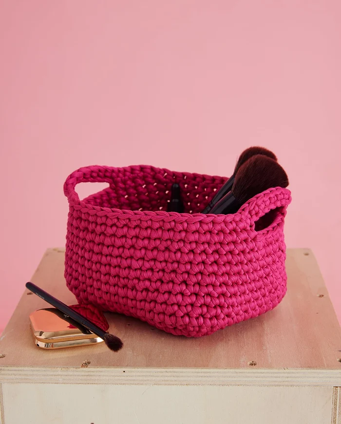 Cesto quadrado rosa vivo trançado com pincéis de maquilhagem, sobre caixa de madeira clara, fundo rosa.