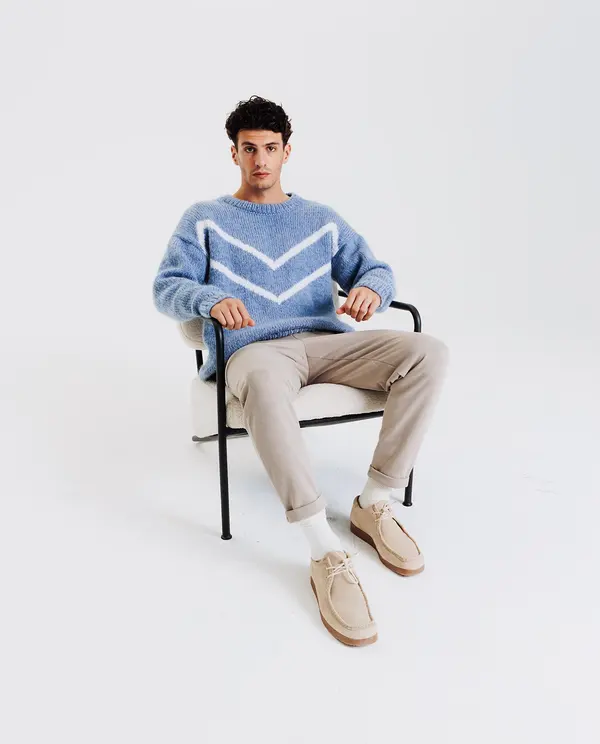 Homem sentado veste camisola azul clara com padrão branco, calças bege, meias e sapatos bege