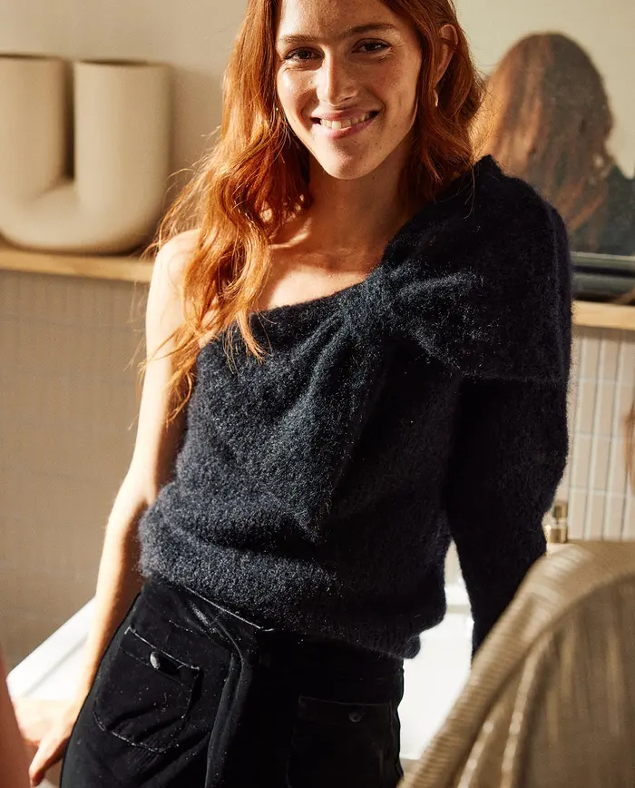 Mulher ruiva a usar top preto felpudo de uma manga só, num ambiente interior com espelho e decoração atrás.