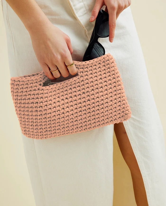 Carteira rosa de crochê com óculos escuros e telemóvel nas mãos, calças brancas com fenda