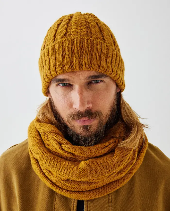 Homem com gorro e cachecol amarelo mostarda de malha e casaco castanho.