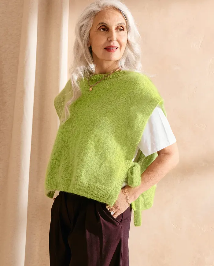 Mulher vestindo colete verde pistácio de malha por cima de t-shirt branca e calças escuras, em fundo bege.